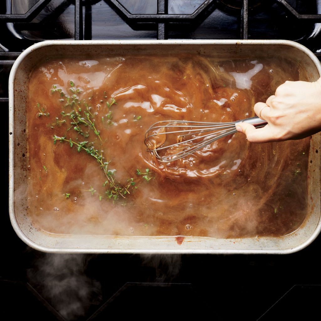 A hand using a whisk to stir a dark gravy in a roasting pan on a stovetop, with sprigs of fresh thyme floating in it