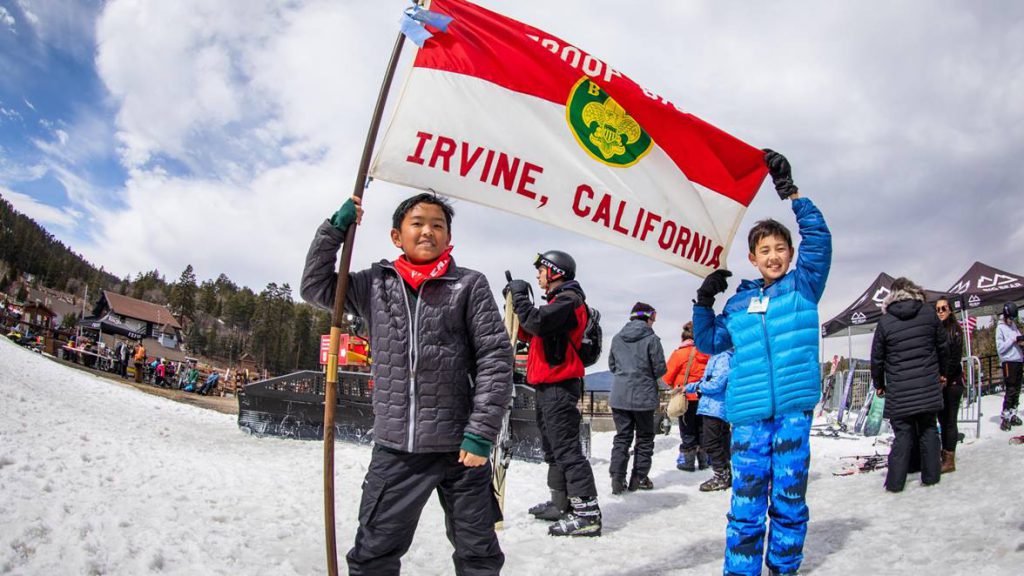 Two children stand in the snow, smiling and holding a large red and white flag that reads "Irvine, California" at Big Bear