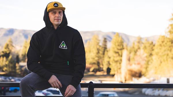 Model wearing a black hoodie and yellow cap with mountain scenery in the background.