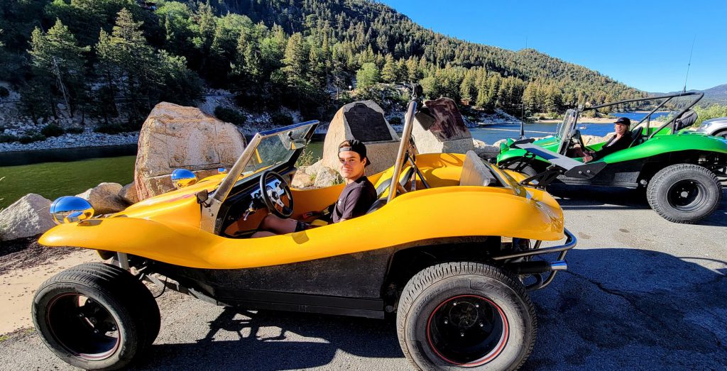 Two people drive green dune buggies on a dirt road, surrounded by pine trees and a calm lake at Big Bear Lake, California.
