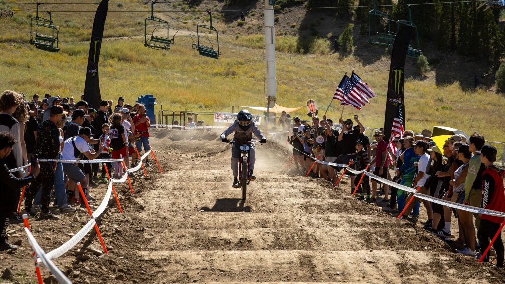 Mountain biker racing downhill during the Monster Energy Pro Downhill Series at Snow Summit in Big Bear, surrounded by cheering crowd.