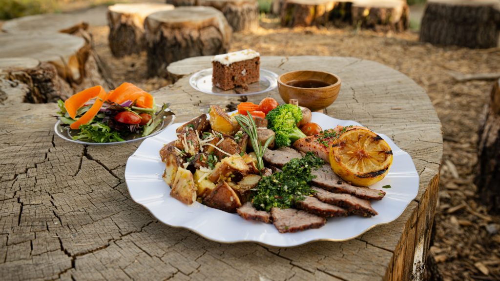 Outdoor meal with plated food and salad on a rustic wooden table at sunset