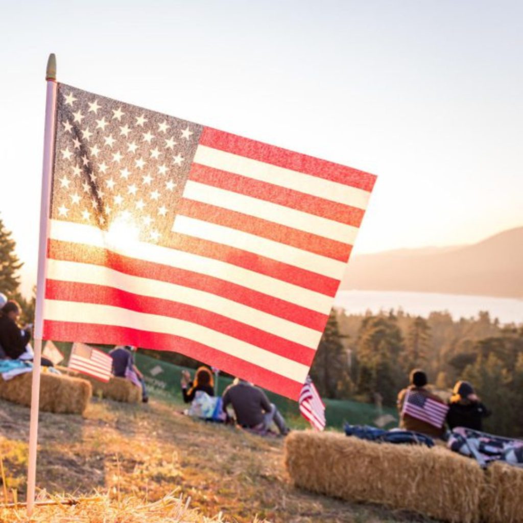 American flag on a pole in the foreground with people sitting on hay bales in the background, scenic outdoor view at sunset