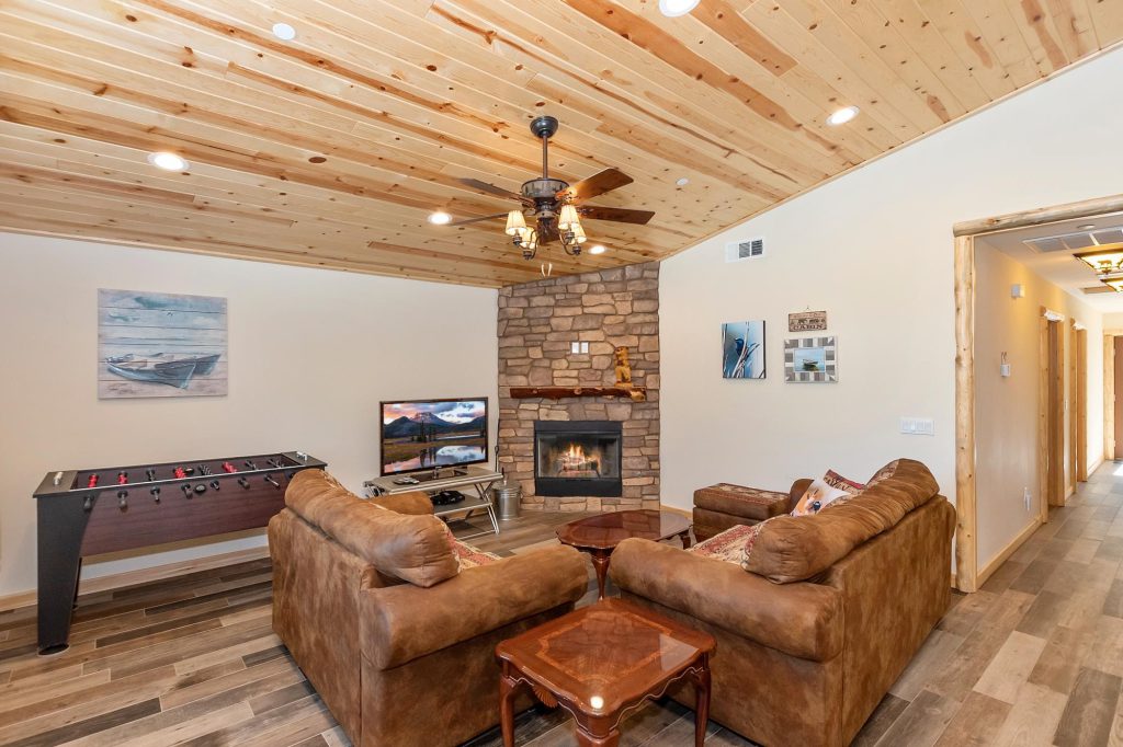 A cozy living room with a stone fireplace, two brown leather couches, a TV, a foosball table, and a wood-paneled vaulted ceiling in a cabin in Big Bear.