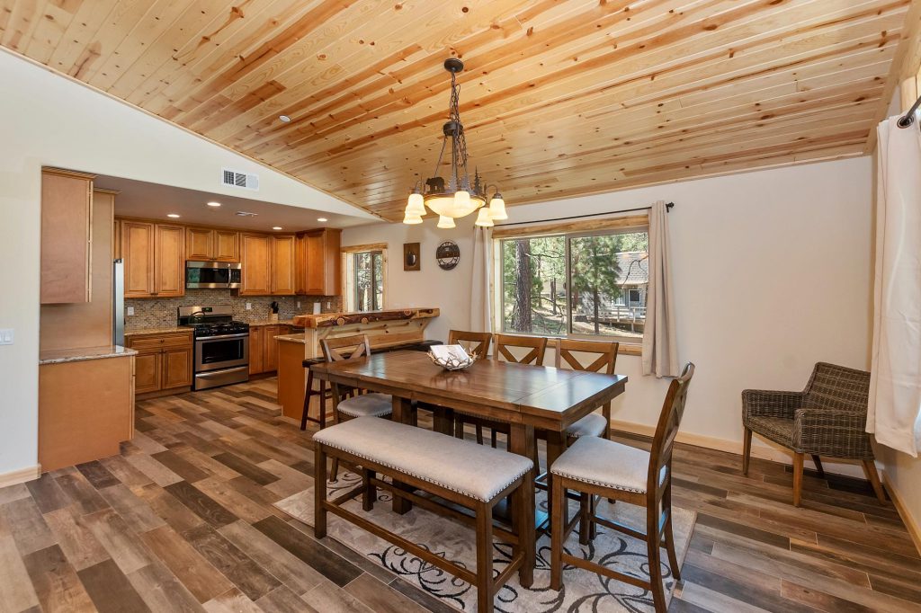 An open-concept dining room with a large wooden table and bench seating, leading into a kitchen with wood cabinets and stainless steel appliances in a cabin in Big Bear.