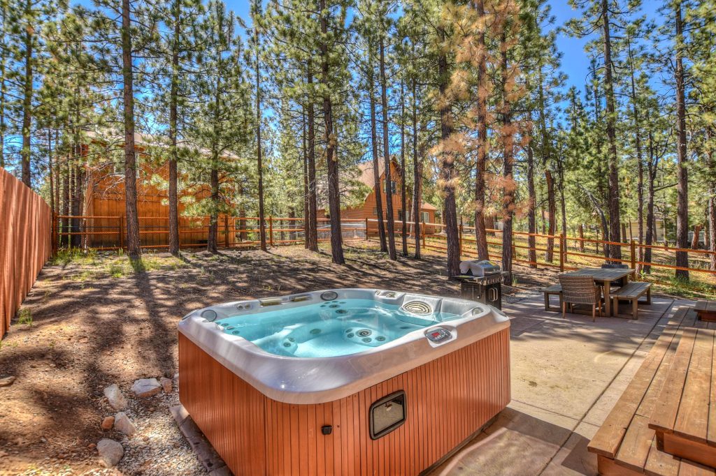 An outdoor hot tub sits on a patio next to a wooden cabin and surrounded by a yard with tall pine trees on a sunny day in Big Bear.
