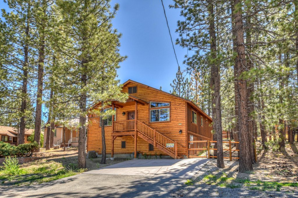 A two-story wooden cabin with a deck and outdoor stairs is surrounded by tall pine trees on a sunny day in Big Bear.