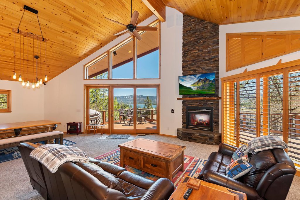 Spacious luxury mountain cabin living room with fireplace, high ceilings, large windows, and scenic lake and forest views from the deck