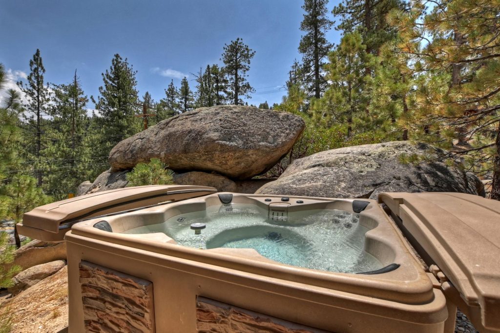 Outdoor hot tub surrounded by large natural boulders and pine trees in Big Bear with scenic mountain surroundings and clear skies.