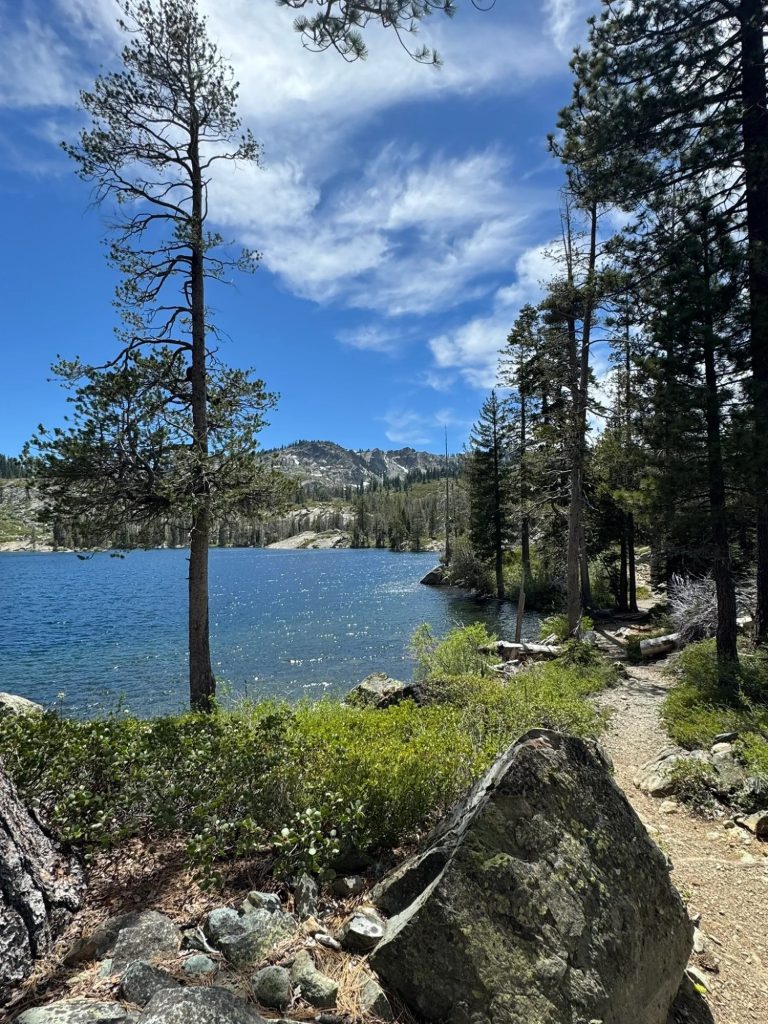 Scenic mountain lake shoreline with a walking trail, pine trees, rocks, and clear blue water under a bright sky with scattered clouds.