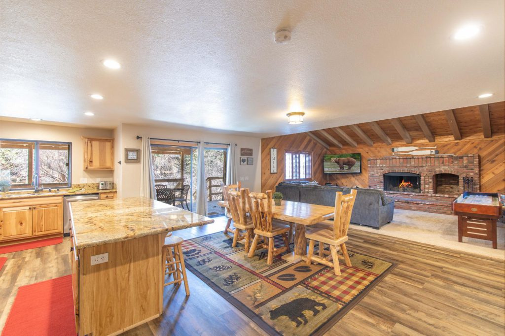 Rustic cabin interior featuring open kitchen, wooden dining table, brick fireplace, and cozy living area