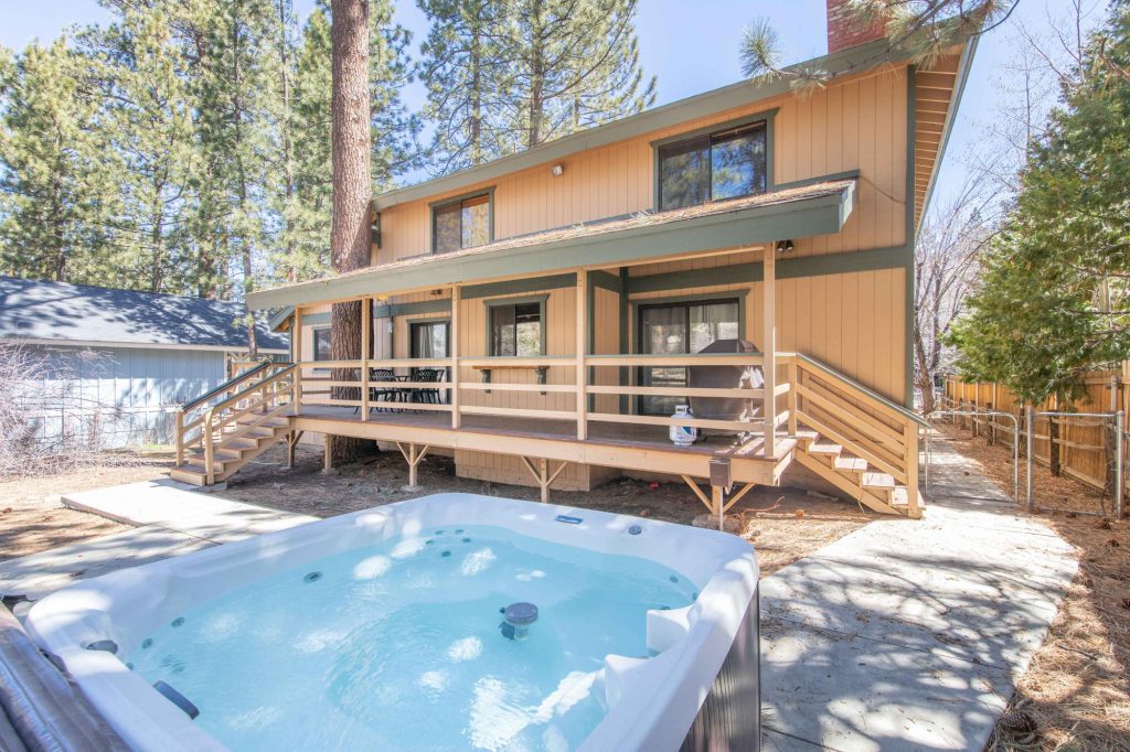 The backyard of a two-story cabin, featuring a hot tub on a concrete patio and a wooden deck with stairs in Big Bear.