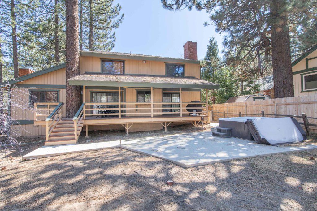 The back of a two-story cabin with a wooden deck, hot tub, and concrete patio in Big Bear.