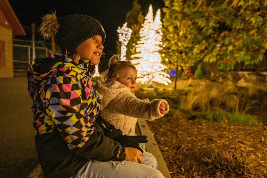 Two small children in winter coats sitting looking at something in the distance with christmass lights on trees behind them. Their faces are glowing and the leaves on the tress are in fall colors