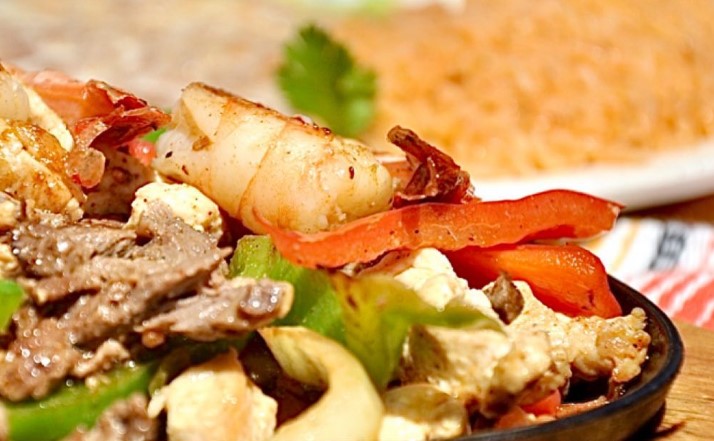 Grilled Shrimp, carne asada, green peppers, red peppers and grilled onions on a black plate with friend Spanish rice in the background