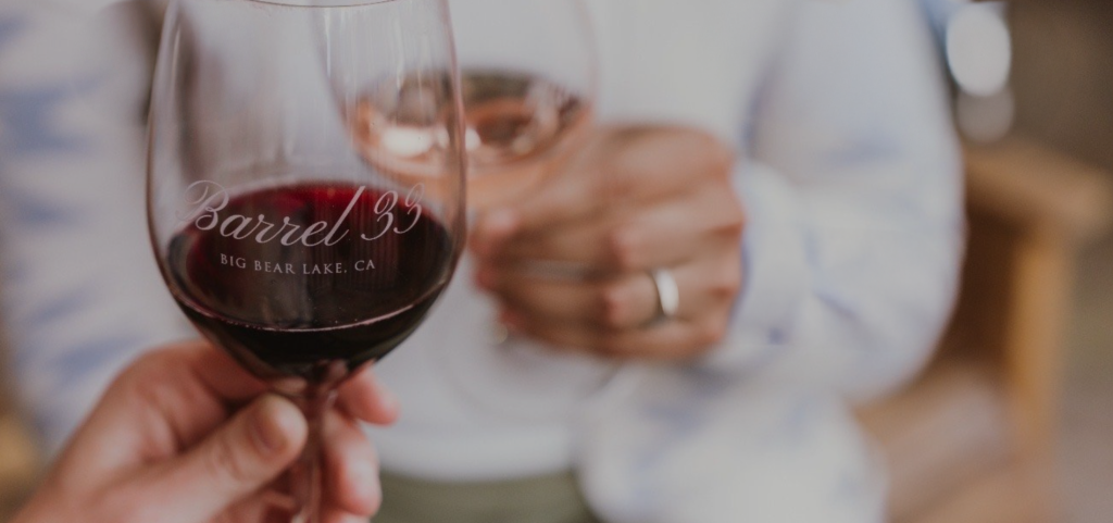 Two people clink glasses of wine, one red and one rosé, with "Barrel 33 Big Bear Lake, CA" etched on the glass in the foreground at Big Bear