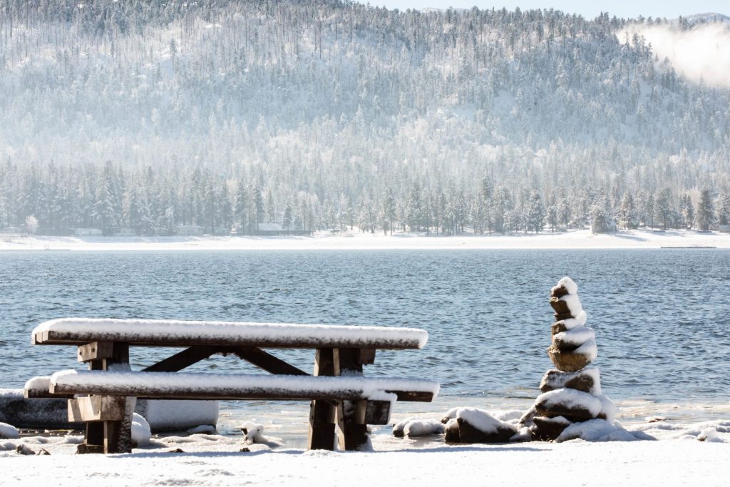 A snow-covered picnic table and a stacked rock sculpture are shown on the snowy shoreline of Big Bear Lake, with a clear blue sky and low-lying clouds on the horizon.