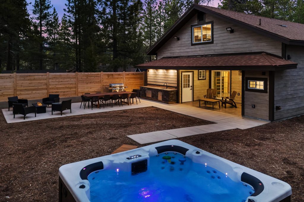 Modern cabin backyard with hot tub, outdoor dining area, patio seating, and fenced yard surrounded by trees at dusk