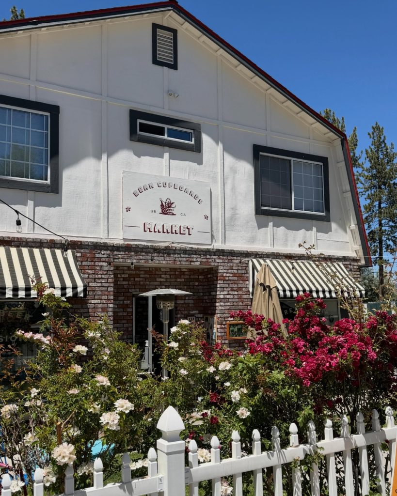 Exterior of Bear Cupboards Market featuring blooming rose gardens, striped awnings, and a white picket fence in Big Bear.