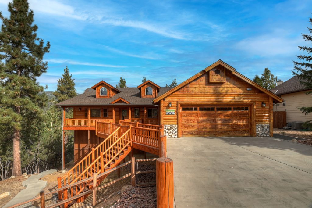 Single level log cabin with a tall set of stairs on the left. Surrounded by pine trees and blue skies.
