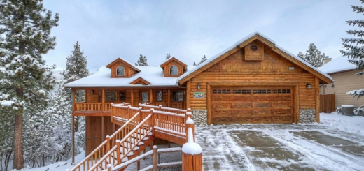 Single level log cabin with a tall set of stairs on the left. Surrounded by pine trees and snow covered.