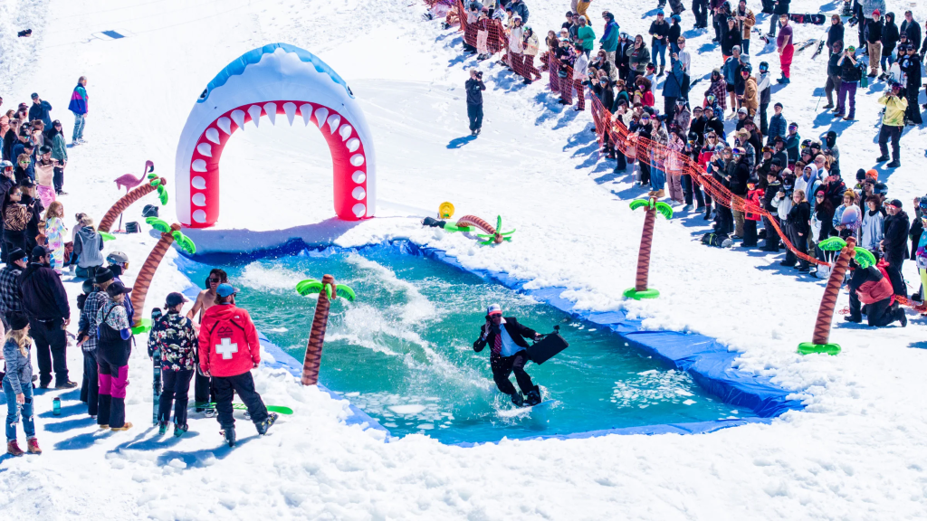 A person in a suit skis across a pond of water at the Bear Break pond skimming event at Big Bear Mountain Resort, with spectators watching from the snowy slope.