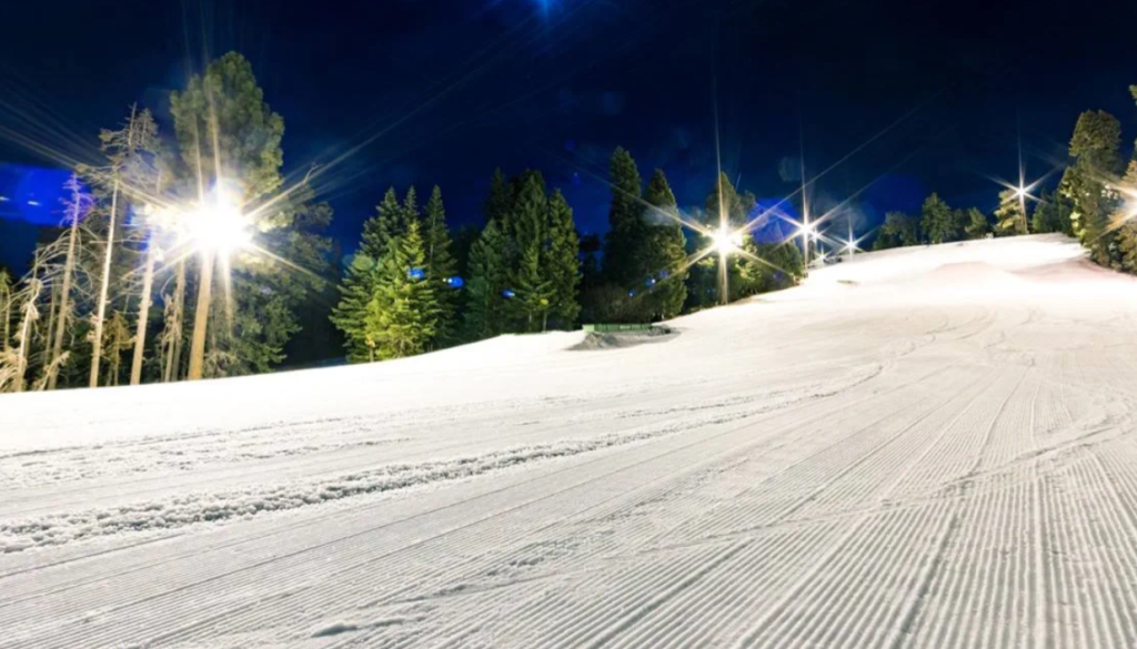 Floodlit ski slope at night at Big Bear Mountain Resort with groomed snow and pine trees in the background.