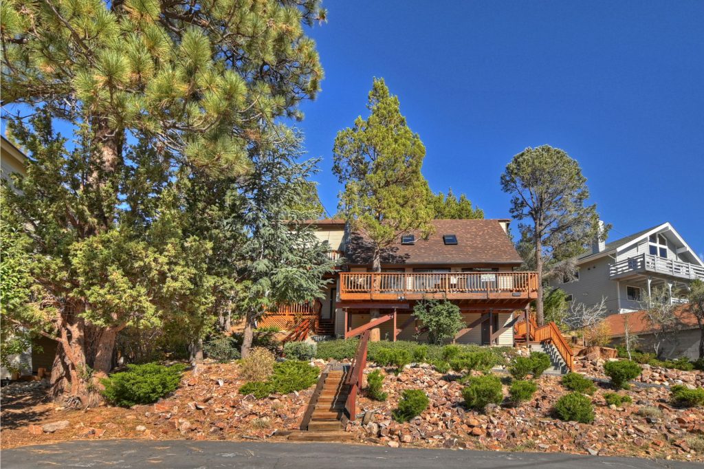 The exterior of a multi-level Big Bear cabin is shown, with a wooden deck, a steep wooden staircase leading up to the front door, and surrounded by large pine trees under a clear blue sky.