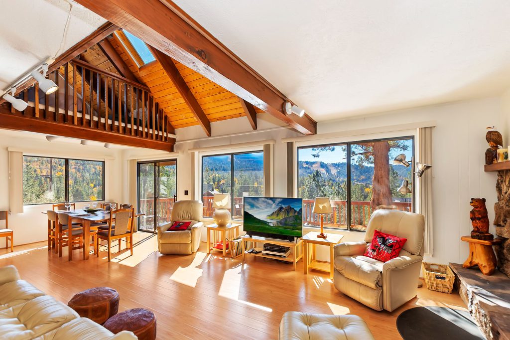 The living room of a Big Bear cabin features high vaulted wooden ceilings with a loft, large windows with a scenic view, and a dining area, with two reclining chairs facing a television.