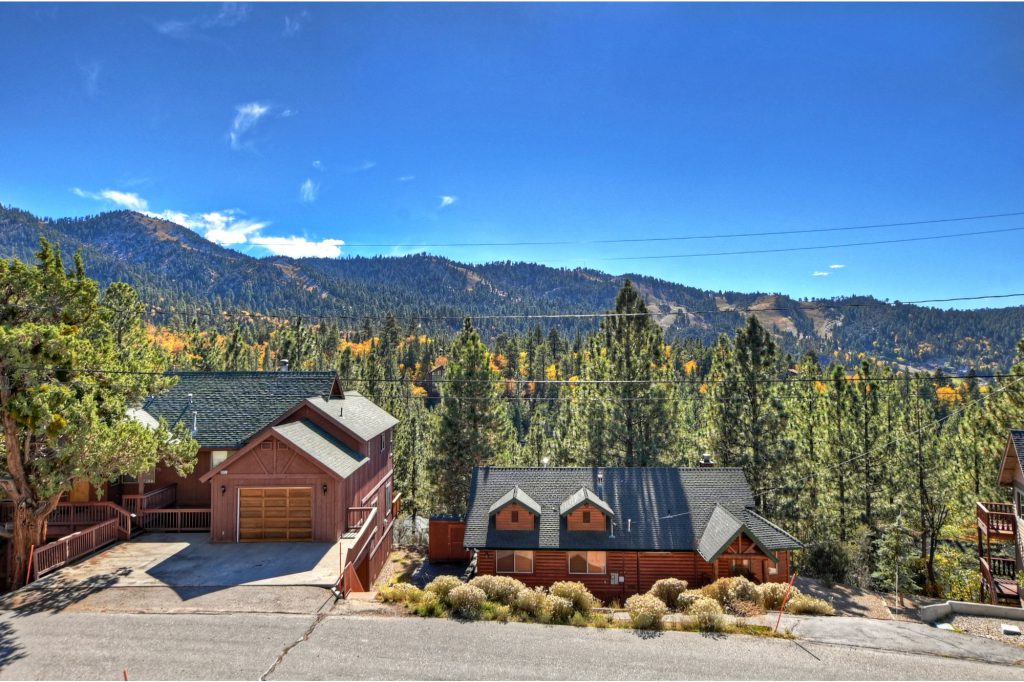 Two wooden cabins are visible on a paved street in Big Bear, with a mountain covered in pine trees and patches of yellow foliage in the background under a bright blue sky.