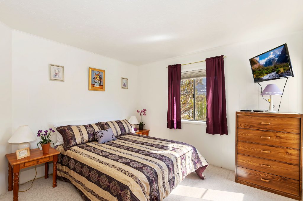 A cozy bedroom in a Big Bear cabin is shown with a bed covered in a patterned comforter, a wooden dresser with a television mounted above it, and a window with purple curtains.