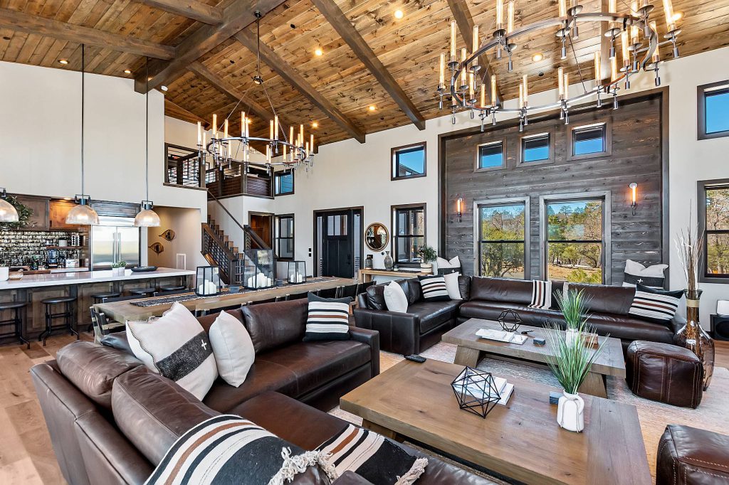 The interior of a modern, open-concept Big Bear lodge with a large living room, a kitchen with a bar, and vaulted ceilings with chandeliers.