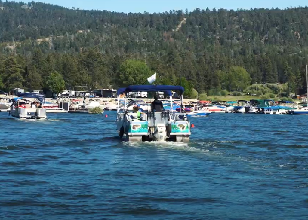 A pontoon boat is cruising on Big Bear Lake, with a marina and forested shoreline visible in the background under a sunny sky.