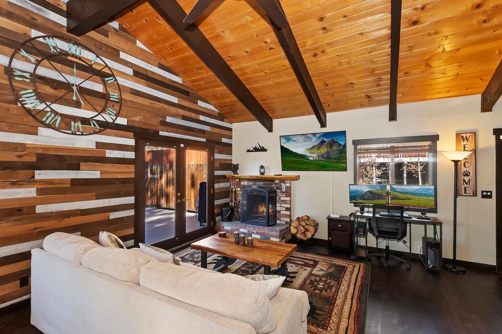 Rustic mountain cabin living room with wood accent walls, stone fireplace, cozy sofa seating, and workspace desk setup with scenic views