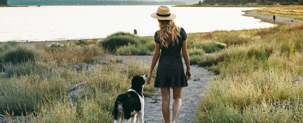 A woman in a sun hat and a dress walks barefoot on a grassy path with a dog by her side, near a body of water in Big Bear.