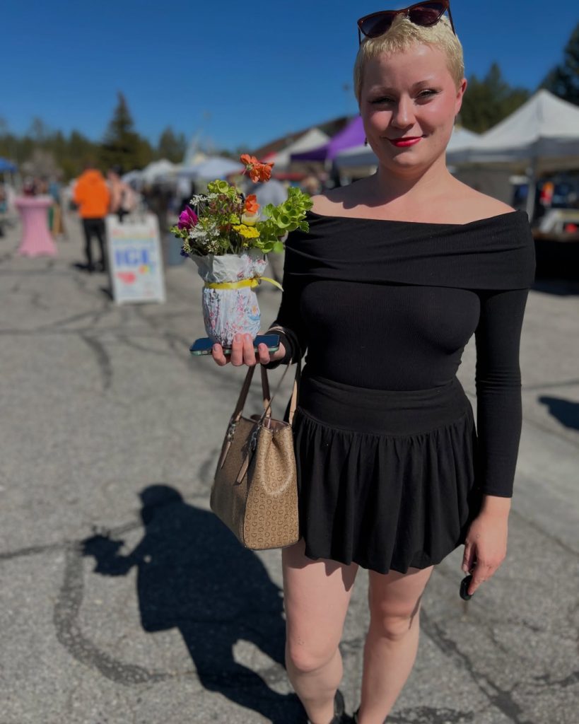 isitor holding a colorful flower bouquet at an outdoor farmers market with vendor tents in the background on a sunny day.