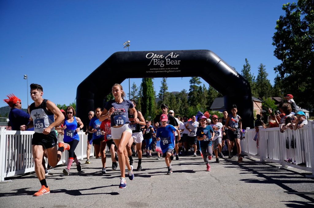 Group of runners starting a road race under a large inflatable “Open Air Big Bear” arch, with participants wearing race bibs and running through a fenced start corridor on a sunny day.