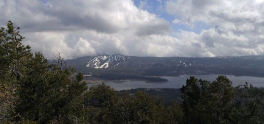 Scenic overlook of Big Bear Lake surrounded by forested mountains, with snow patches on distant peaks and dramatic clouds over the landscape.