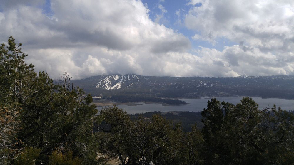 Scenic overlook of Big Bear Lake surrounded by forested mountains, with snow patches on distant peaks and dramatic clouds over the landscape.