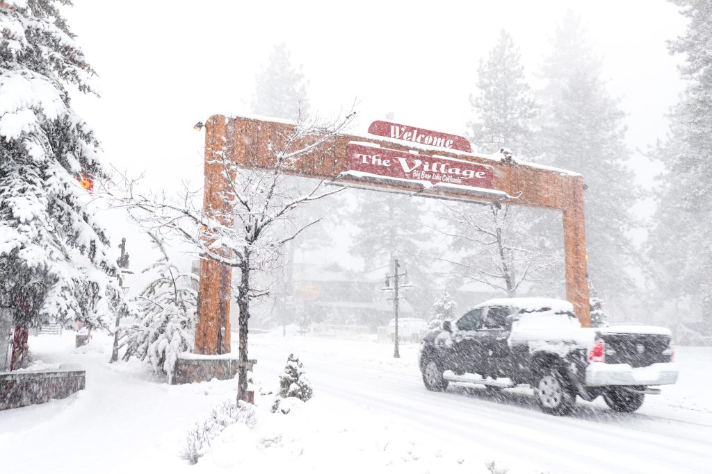 A heavy snowfall is shown in Big Bear, with a snow-covered truck driving under the large "Welcome to The Village" sign.
