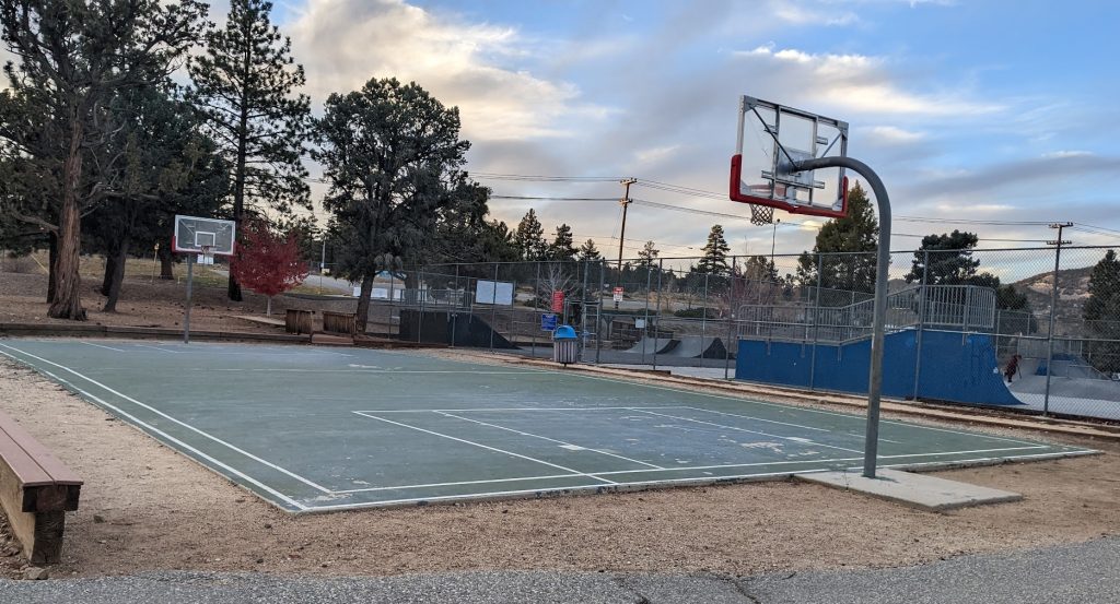 Outdoor basketball court in a mountain park setting with surrounding trees, skate park area, and a cloudy sky in the background.