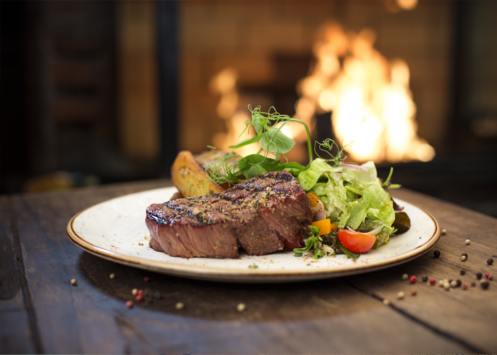 A grilled steak with a side salad and potatoes is presented on a plate in a cozy Big Bear setting, with a warm fireplace blurred in the background.