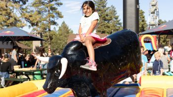 Young girl smiling while riding a mechanical bull at an outdoor fair with people and tents in the background