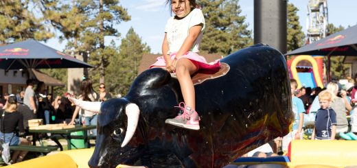 Young girl smiling while riding a mechanical bull at an outdoor fair with people and tents in the background