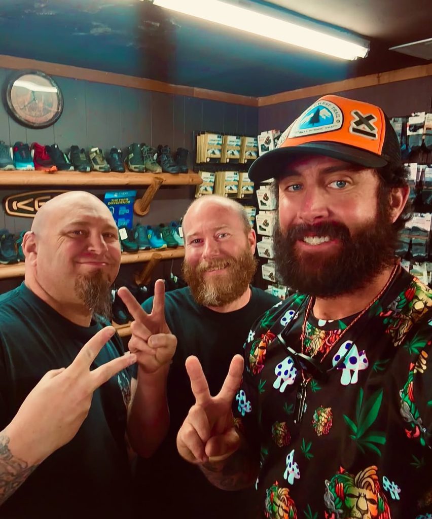 Three men posing inside a sporting goods or bike shop, smiling and showing peace signs, with shelves of cycling shoes and outdoor gear in the background.