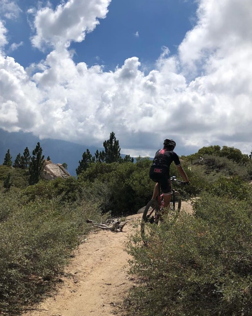 A mountain biker rides along a narrow singletrack trail surrounded by shrubs, boulders, and mountain scenery in Big Bear.