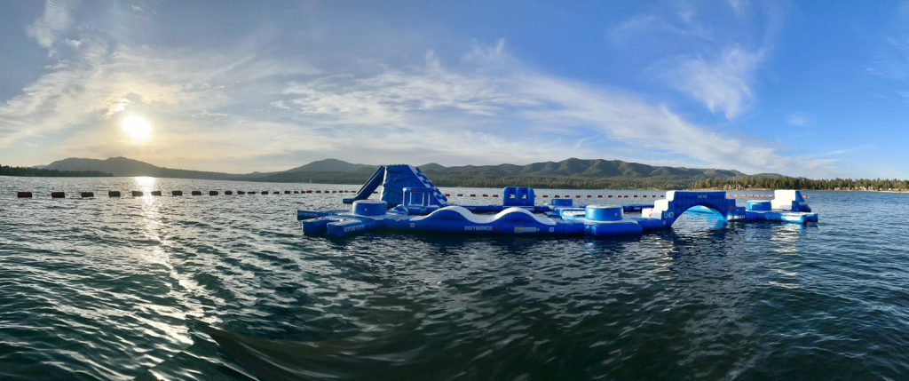A wide shot shows a large blue inflatable waterpark floating on the calm waters of Big Bear Lake under a sunny sky.