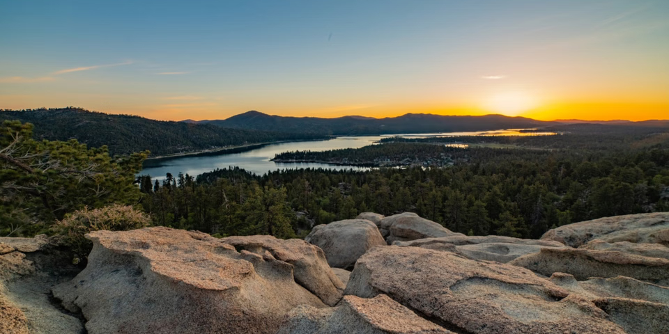 Panoramic sunset view overlooking Big Bear Lake from a rocky viewpoint, with forested mountains and the lake stretching into the distance.
