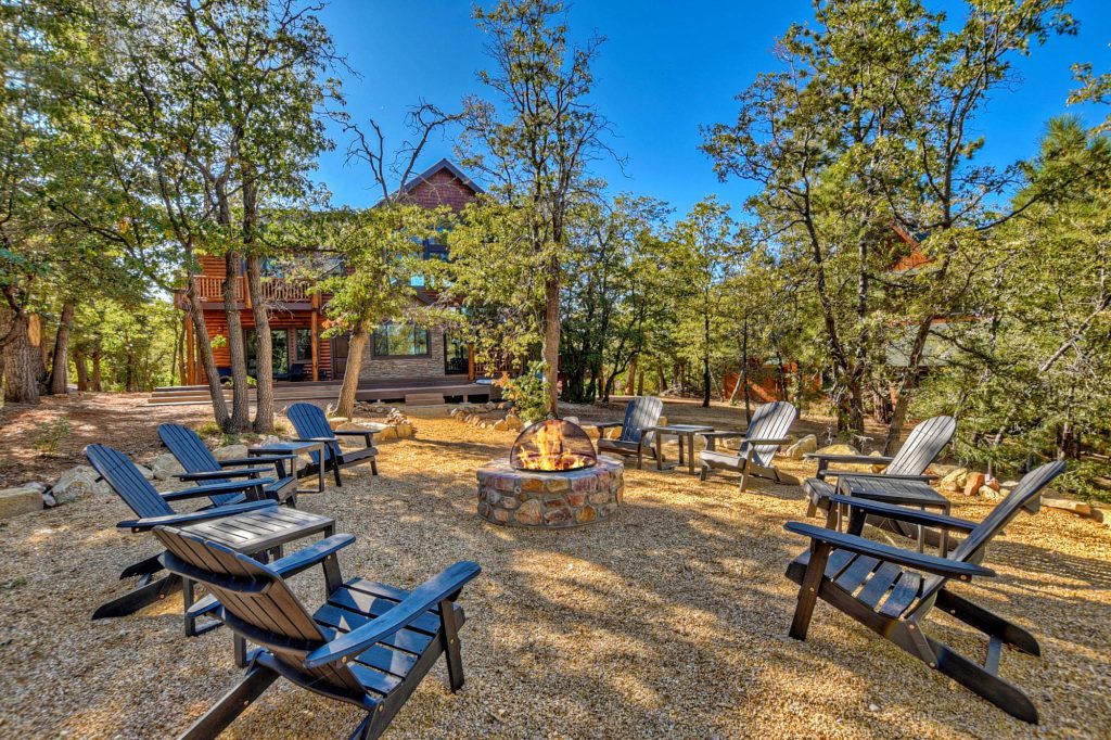A rustic fire pit with Adirondack chairs is set up in a wooded area in front of a multi-story cabin, with trees and blue skies visible at Big Bear.