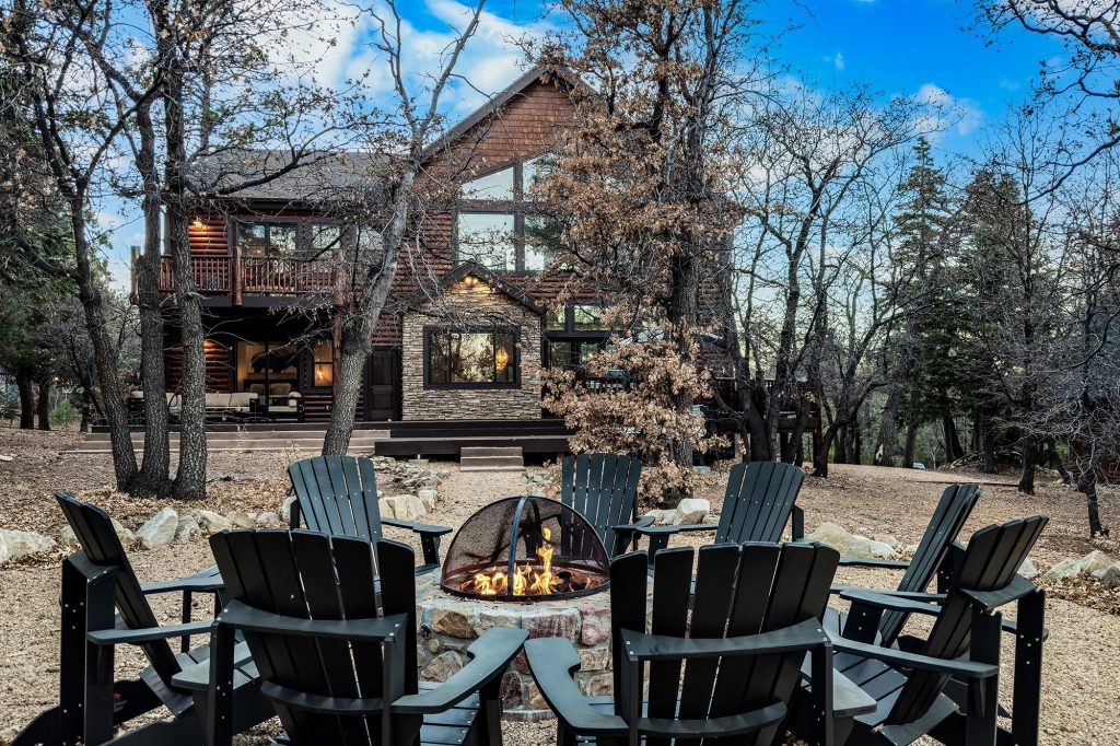 The exterior of a large rustic wooden cabin in Big Bear, with a circular arrangement of black chairs around a fire pit in the foreground.